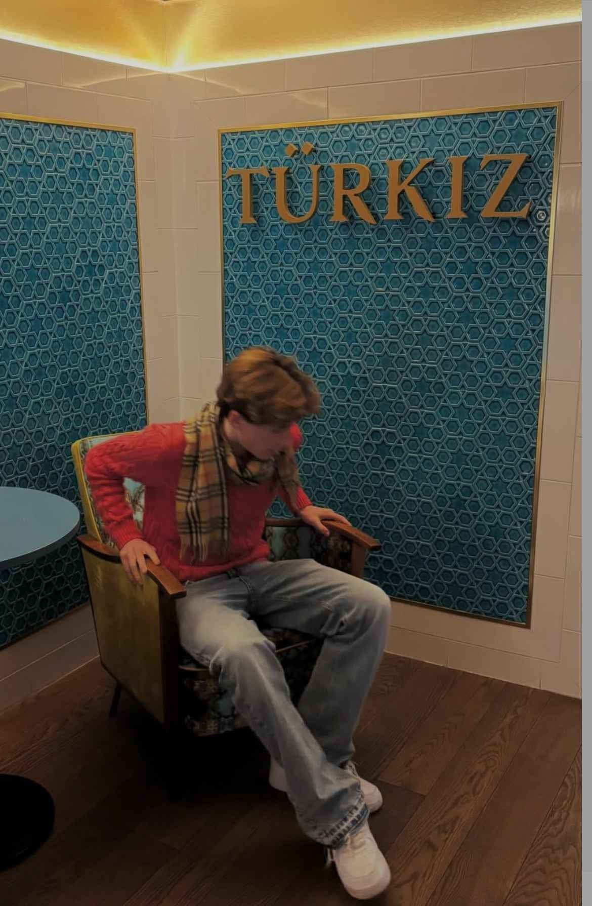 Fiatal férfi streetwear stílusban farmernadrágban, piros pulóverben, sálban és fehér cipőben ül egy vintage székben, modern, kék mozaikos fal előtt, háttérben 'TÜRKIZ' felirat.