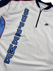 Fehér Nike streetwear felső, SUPREME felirattal, kontrasztos kék-piros részletekkel.